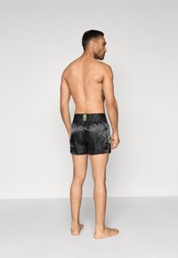 Melnas satīna peldbikses ar elastīgu jostu, kas ir rotātas ar zaļu logotipa elementu un gludu tekstūru. Dizainētas ciešai pieguļošai izskatam.