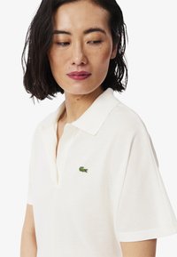 Vit polo skjorta med en texturerad finish, med en grön broderad krokodil-logotyp på vänster bröst. Klassisk krage och korta ärmar.