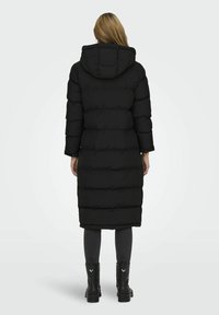 Lång svart pufferjacka med huva, med en quiltad design, längd till mitten av vaderna, och framfickor. Bärs med grå leggings och svarta stövlar.