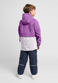 Paarse en witte waterdichte jas met een capuchon, met elastische manchetten en reflecterende accenten, gecombineerd met marineblauwe broek en witte schoenen.