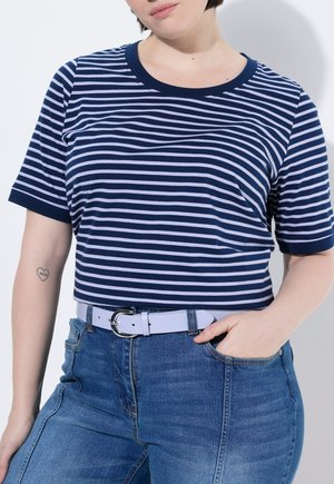 Femme portant un t-shirt rayé bleu marine et blanc, rentré dans un jean bleu taille haute avec une ceinture blanche, main dans la poche, tatouage en forme de cœur sur le bras.