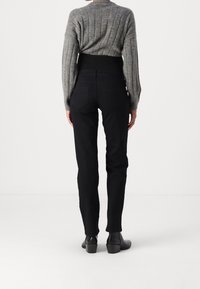 Persona que lleva vaqueros negros de talle alto, suéter gris con textura de canalé y botas de tobillo negras con tacón, de pie de espaldas sobre un fondo blanco liso.