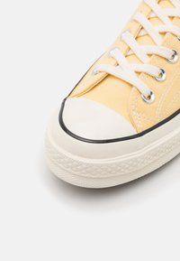 Baskets en toile jaunes avec lacets blancs, passepoil noir et capuchon en caoutchouc. La semelle texturée présente un motif en diamant pour une meilleure adhérence.