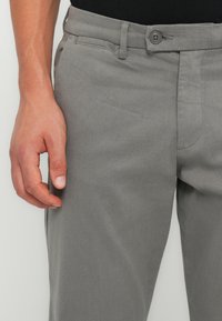 Pantaloni grigi in misto cotone con una texture liscia, dotati di chiusura con bottone, tasche frontali e design slim fit. Mano posizionata di lato.