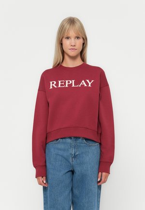 Felpa rossa con la scritta "REPLAY" stampata in bianco sul petto. Presenta un collo rotondo e una vestibilità comoda, abbinata a jeans azzurri chiari.