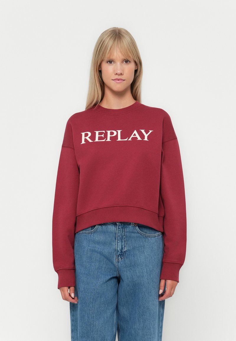 Sweatshirt rouge avec "REPLAY" imprimé en blanc sur la poitrine. Doté d'un col rond et d'une coupe décontractée, associé à un jean bleu clair.