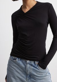 Top nero a maniche lunghe con un design avvolgente, caratterizzato da una texture arricciata su un lato. Abbinato a jeans di denim blu chiaro.