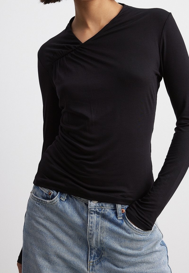 Top nero a maniche lunghe con un design avvolgente, caratterizzato da una texture arricciata su un lato. Abbinato a jeans di denim blu chiaro.