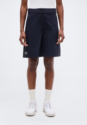 Osoba ubrana w granatowe szorty do kolan z logo EA7 na lewej nogawce, białe skarpetki, białe trampki i biały top, stojąca na jednolitym tle.