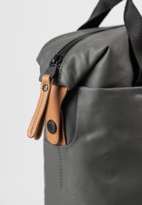 Grijze waterdichte tas met een gladde textuur, voorzien van een zwarte rits en een bruine leren ritslus. Compact ontwerp met een detail van een extern zakje.