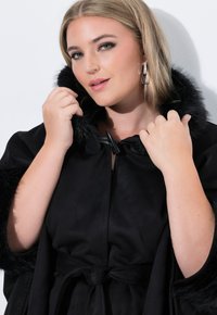 Capa negra con ribete de pelo sintético, con un diseño elegante y una textura suave. Cerrada con un botón de toggle negro en el cuello.