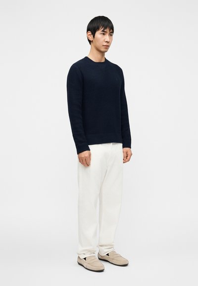 Μπλε-σκούρο πλεκτό πουλόβερ με στρογγυλή λαιμόκοψη, συνδυασμένο με λευκό χαλαρό παντελόνι και ανοιχτό μπεζ παπούτσια χωρίς κορδόνια.