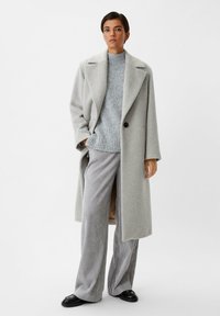 Lichtgrijze oversized jas met geknipte revers, gedragen over een grijze trui en gestreepte wijde broek. Soepele stof met een relaxte pasvorm.