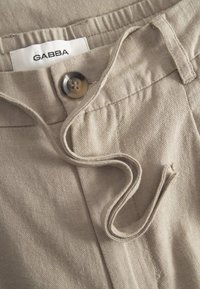 Smėlinės kelnės su minkšta tekstūra, užsegamos su saga, su raišteliu juosmens srityje ir etikete su užrašu „GABBA“ ant juosmens.