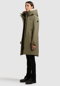 Olivengrøn parka med hætte foret med pels, med lynlåslommer og trykknapsmanchetter, parret med sorte tætsiddende bukser og ankelstøvler.