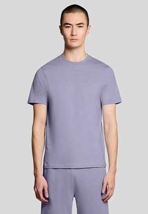 Uomo che indossa una maglietta a maniche corte color viola chiaro e pantaloncini abbinati, in piedi davanti a uno sfondo grigio.