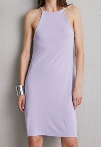 Vrouw in een mouwloze lila jurk met halterhalslijn en zilveren armband, staand tegen een gestructureerde grijze muur.