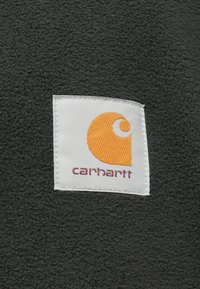 Tessuto di pile nero con un'etichetta bianca quadrata che presenta un logo arancione e il testo "carhartt" in viola sotto il logo.