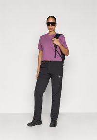 Μωβ κοντομάνικο cropped μπλουζάκι, μαύρο ελαφρύ παντελόνι με λογότυπο, μαύρα γυαλιά ηλίου και ανθεκτικά μαύρα παπούτσια. Σακίδιο πλάτης στον έναν ώμο.