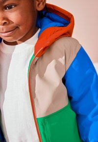 Chaqueta colorida con capucha naranja, mangas azules, frente beige y bolsillo verde. Fabricada con material sintético suave, cuenta con cierre de cremallera.