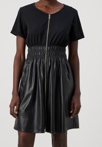 Schwarzes Kleid mit kurzen Ärmeln, elastischer Taille, Reißverschluss und Taschen. Der Stoff ist eine Mischung aus Baumwolle und Kunstleder, glatte Textur.