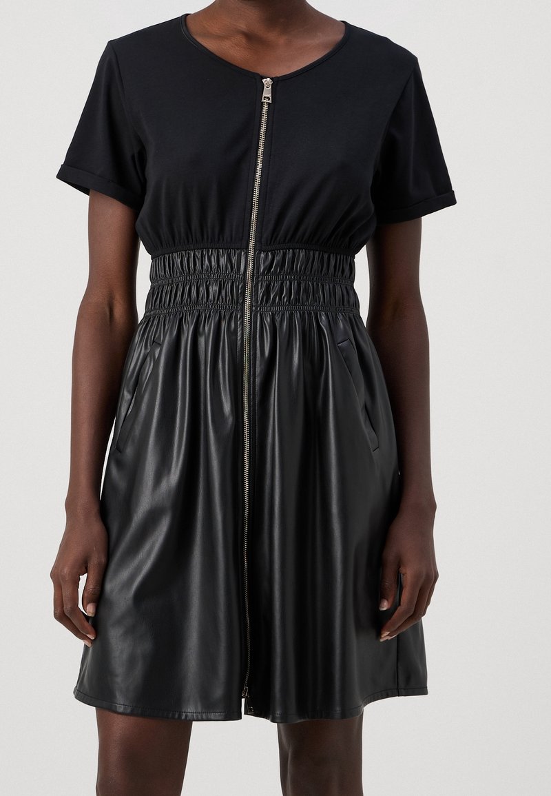 Schwarzes Kleid mit kurzen Ärmeln, elastischer Taille, Reißverschluss und Taschen. Der Stoff ist eine Mischung aus Baumwolle und Kunstleder, glatte Textur.