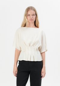 Blusa color crema con maniche corte e ampie, caratterizzata da delle pieghe in vita e un tessuto liscio e strutturato. Abbinata a pantaloni neri.