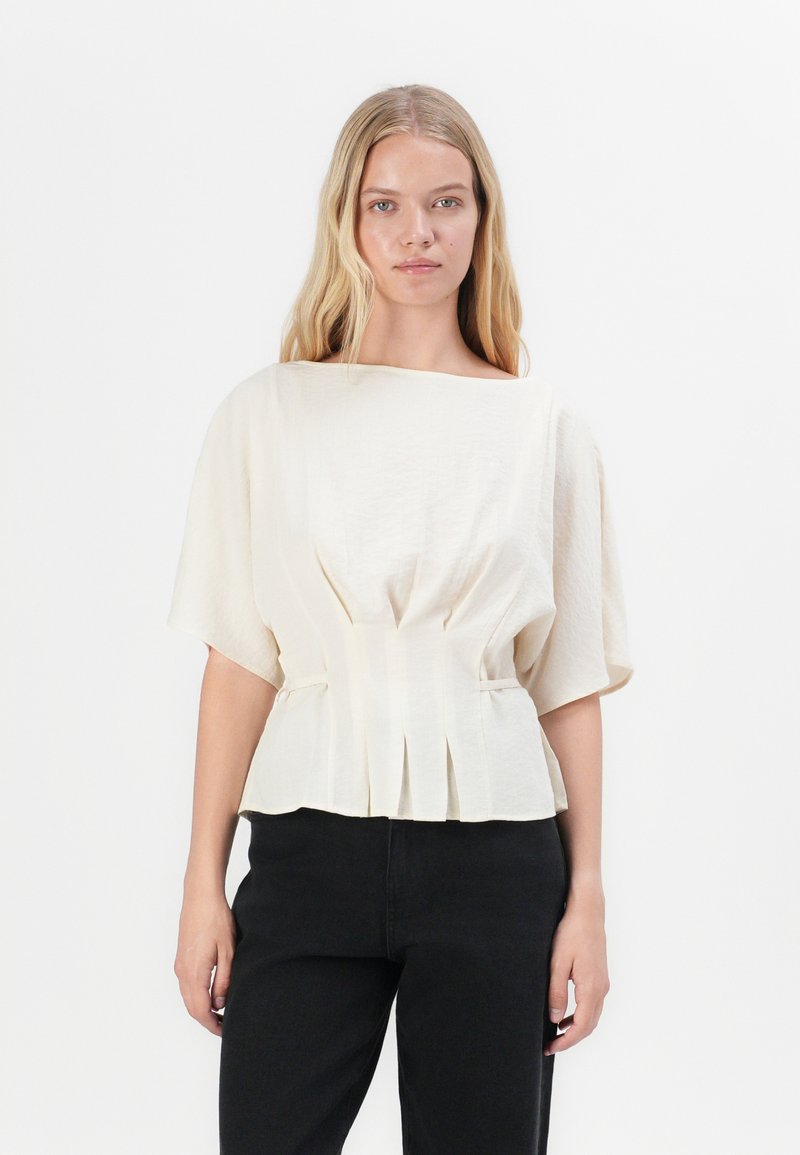 Blusa color crema con maniche corte e ampie, caratterizzata da delle pieghe in vita e un tessuto liscio e strutturato. Abbinata a pantaloni neri.