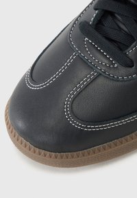 Svart lær sneakers med hvite sømmer, avrundet tå, og gummisåle med strukturert mønster. Snøringen er svart og gir en sikker passform.