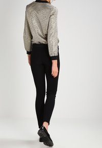 Donna che indossa un top di paillettes argento, pantaloni neri attillati e scarpe nere, in piedi contro uno sfondo chiaro e neutro.