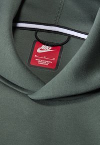 Zelená mikina Nike s jemnou texturou, skládacím límcem a černobílým šňůrkou. Na štítku je uvedena velikost M, vyrobeno v Kambodži.