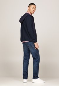 Felpa con zip di colore blu navy con rivestimento viola, indossata sopra una maglietta rosa chiaro e jeans blu. Il modello sta in piedi con le mani in tasca, indossando scarpe bianche.