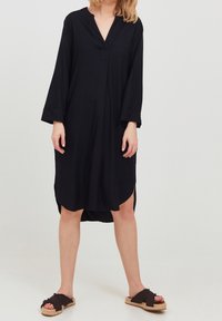 Robe noire midi avec un décolleté en V, manches longues, fentes latérales, fabriquée en tissu lisse. Associée à des sandales sombres avec une lanière texturée.