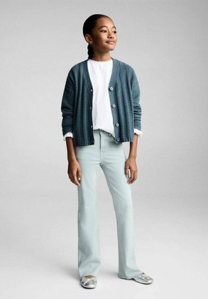 Gilet en tricot turquoise sur une chemise blanche, associé à un jean évasé vert clair et des ballerines argentées. Design simple et texturé avec des boutons.