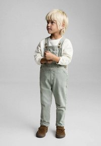Zeleni denim kombinezon s bež gumbi, uparen s belom majico z dolgimi rokavi. Otrok nosi rjave natikače in stoji ob sivi ozadju.