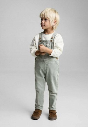 Gröna denimoverall med bruna knappar, i kombination med en vit långärmad skjorta. Barnet har på sig bruna slip-on skor och står mot en grå bakgrund.
