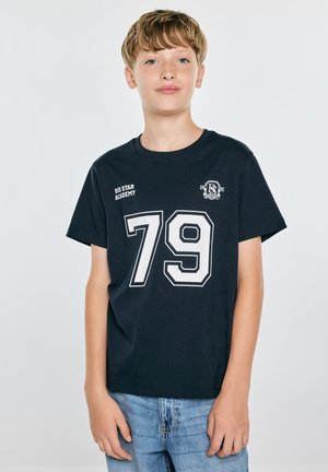 Chłopiec z jasnobrązowymi włosami, ubrany w czarną koszulkę z numerem 79 i tekstem "Big Star Academy,", stojący na jednolitym jasnym tle.