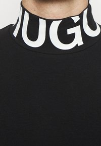 Schwarzes Hemd mit hohem Kragen, das ein auffälliges weißes "HUGO"-Logo um den Kragen trägt, gefertigt aus glattem, elastischem Stoff.