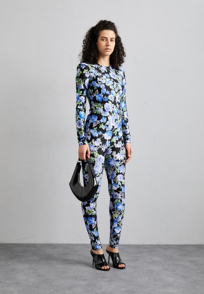 Long-sleeve jumpsuit in black with a vibrant floral pattern in blue and green. Paired with black open-toe heels and a black handbag.