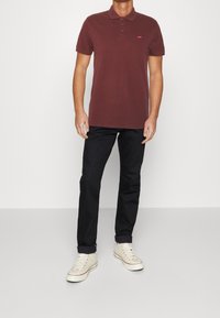 Camisa polo color burdeos hecha de algodón, con una tapeta de tres botones y un pequeño logo rojo. Combinada con pantalones negros y zapatillas beige.