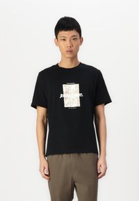 Sort bomulds t-shirt med et centralt grafisk design i beige. Korte ærmer og rund halsudskæring. Bæres med lysebrune bukser.