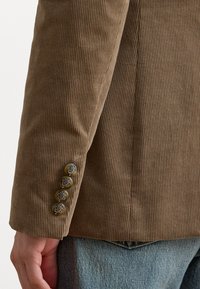 Manica di un blazer in velluto a coste marrone, con una sottile trama a coste, decorata con quattro bottoni in finitura argento vicino al polsino.