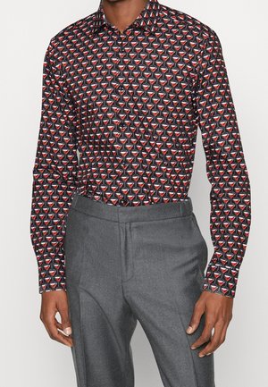 Homme portant une chemise noire à manches longues boutonnée avec un motif de verres à cocktail rouge et blanc et un pantalon gris.