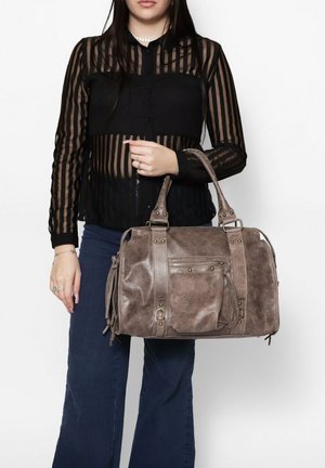Borsa a tracolla grigia con texture, manici doppi, tasca frontale con zip e accessori decorativi. Indossata da una persona con una blusa trasparente a righe nere.