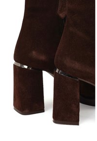 Botas de tacón alto de ante marrón con puntera cuadrada y tacones gruesos en bloque que presentan detalles metálicos donde el tacón se une con la parte superior.