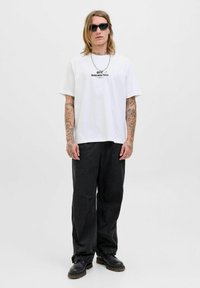 Ung person med langt hår og tatoveringer iført sorte solbriller, hvid UFC Undeniable Force T-shirt, løse sorte bukser og sorte støvler.