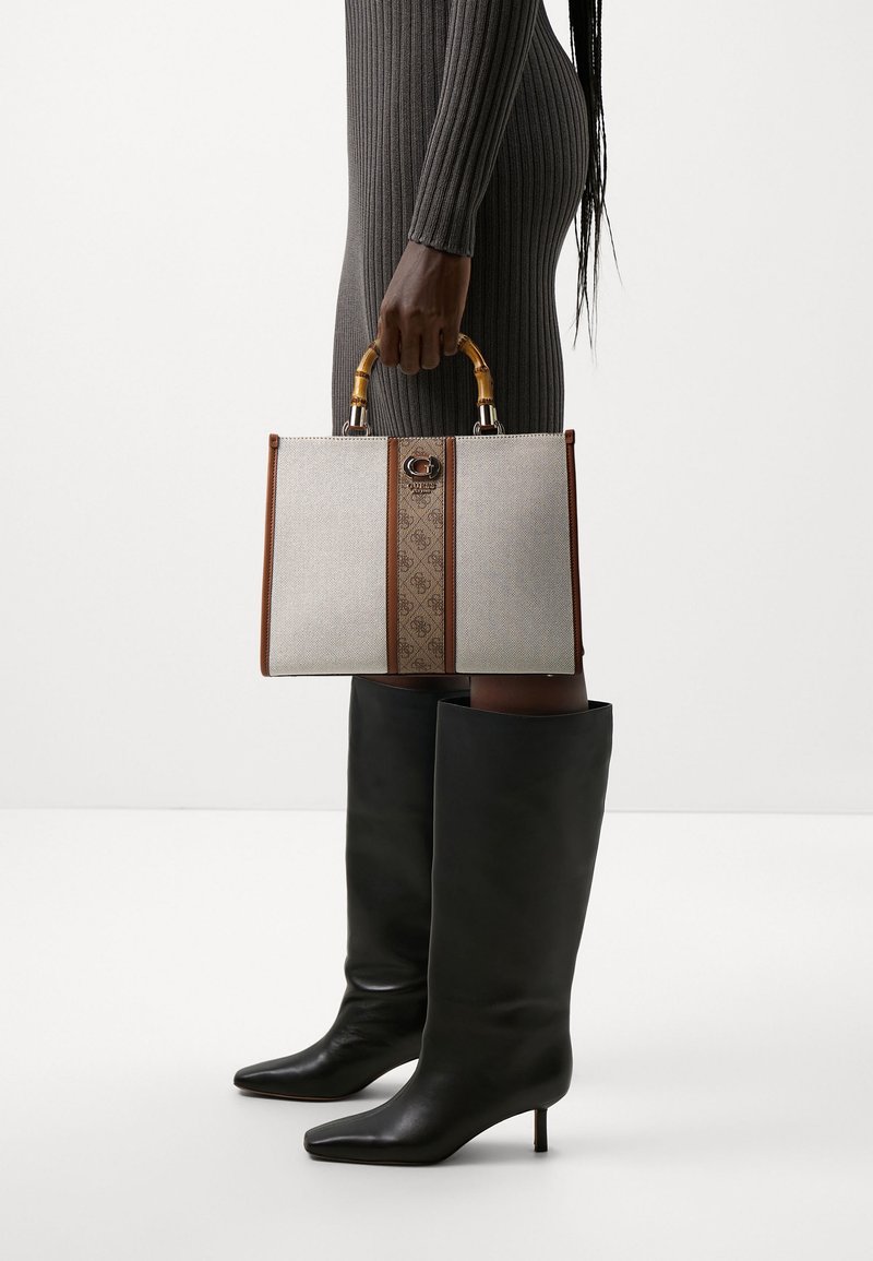 Personne portant une robe sombre côtelée et des bottes noires hautes jusqu'aux genoux, tenant un sac à main structuré beige et marron avec des poignées en bambou.