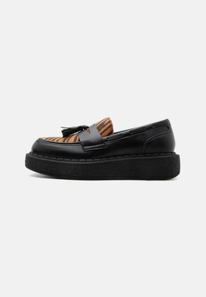 Mocasín plataforma negro con parte superior texturizada con rayas de tigre y borla de cuero negra, sobre un fondo liso gris claro.