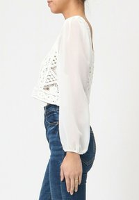 Crop top blanc avec un design en crochet en dentelle à l'avant et des manches longues transparentes. Associé à un jean en denim bleu.