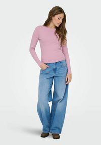 Frau mit langen braunen Haaren, die ein pinkes langärmliges Oberteil, weite hellblaue Jeans und braune Schuhe trägt, steht mit einer Hand in der Tasche.